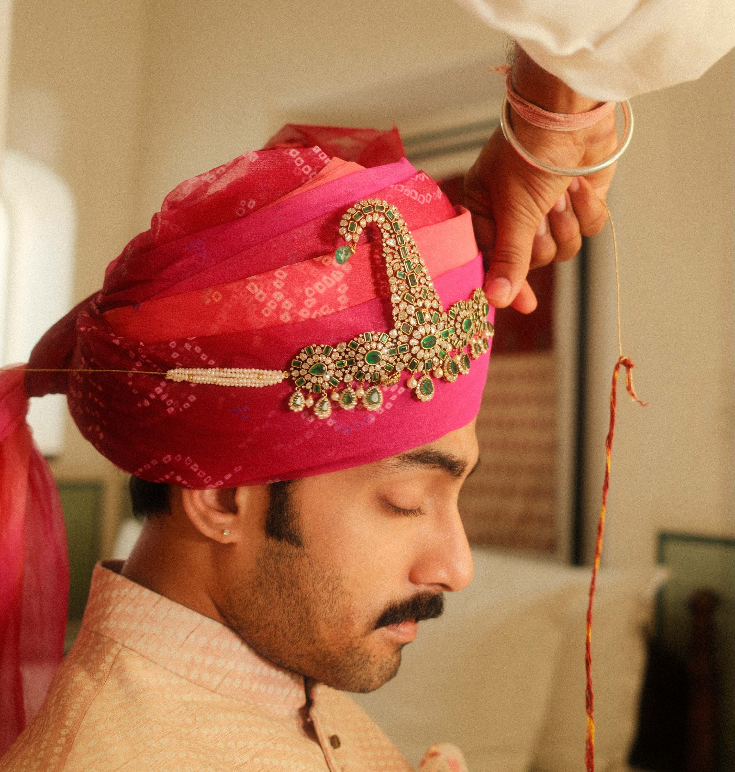 A gold and emerald sarpech being pinned to a groom's pink safa